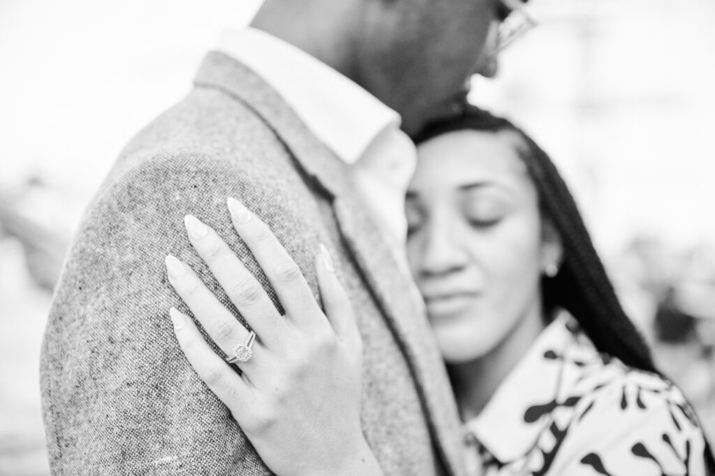 Black & White photo showing Couple in a loving embrace after proposal in NYC at Pier 17 Rooftop. Image shows closeup of engagement ring.