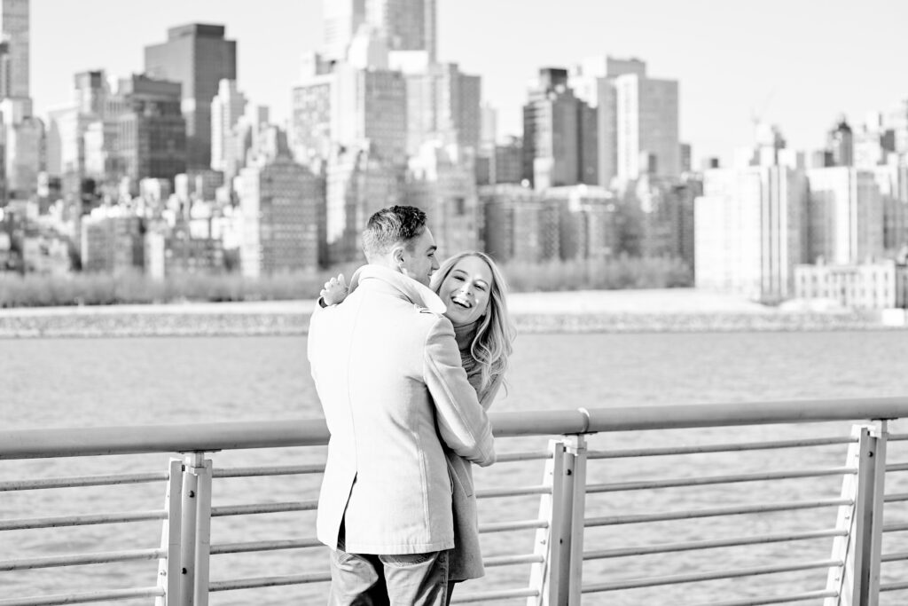 Black and white image of a couple in Gantry Plaza Park shortly after a proposal.  Behind couple is the NYC skyline from the east river. Photo taken by Corey Lamar Photography