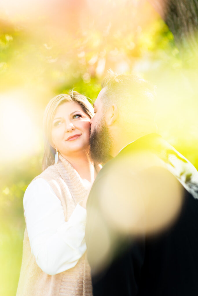Endearing Kiss given on the cheek, a loving couple is depicted within the image.  Foliage is used in front of the camera lens to create a beautiful effect.