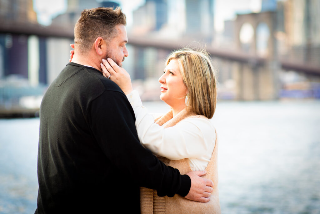 Couple stand tenderly holding each other along the water, in view you can see the Brooklyn Bridge as well as a portion of the NYC skyline