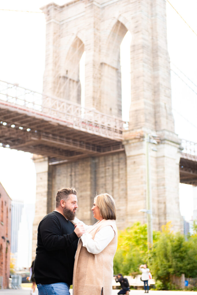 Brooklyn Bridge Appears in the Background as a couple stand together in an embrace looking endearingly at each other. Captured by Corey Lamar Photography
