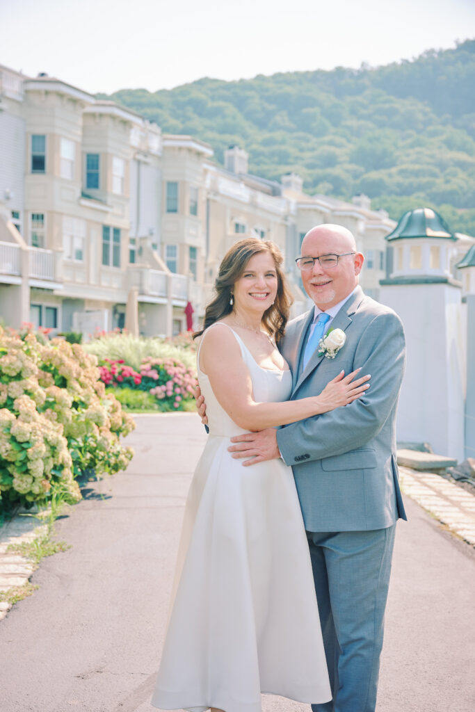 Image of Bride and Groom, they stand embracing in front of their small village in Harbor Pointe, Harverstraw NY in the Lower Hudson Valley