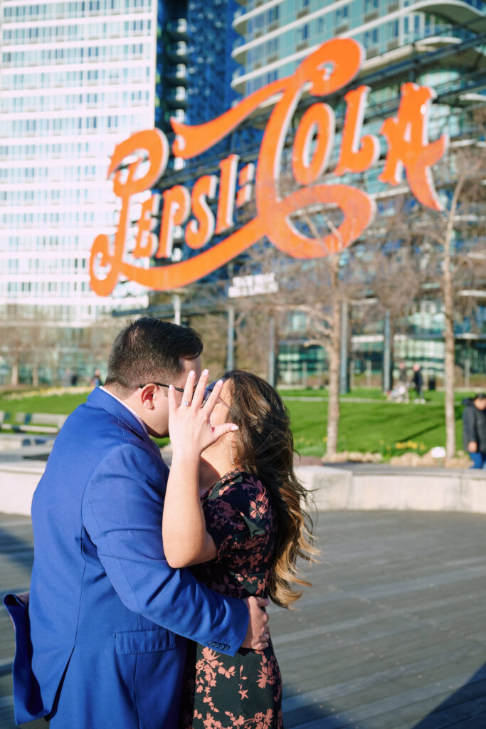 Chris and Katherine kiss, Katherine blocks their face in an artful style showing off her ring.  They stand in front of the Iconic Pepsi Cola Sign in New York City.