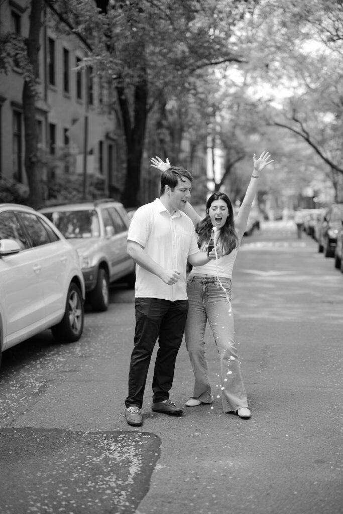 Black-and-white West Village engagement photo of Mark and Vanessa popping champagne in the street